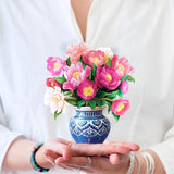 A person in a white shirt is holding the FreshCut Paper's Mini Peony Paradise, showcasing vibrant pink and white flowers. The person's head is not visible, and they are adorned with bracelets on both wrists. The attention is captivated by the meticulously detailed and colorful flower arrangement that exudes classic sentiments.