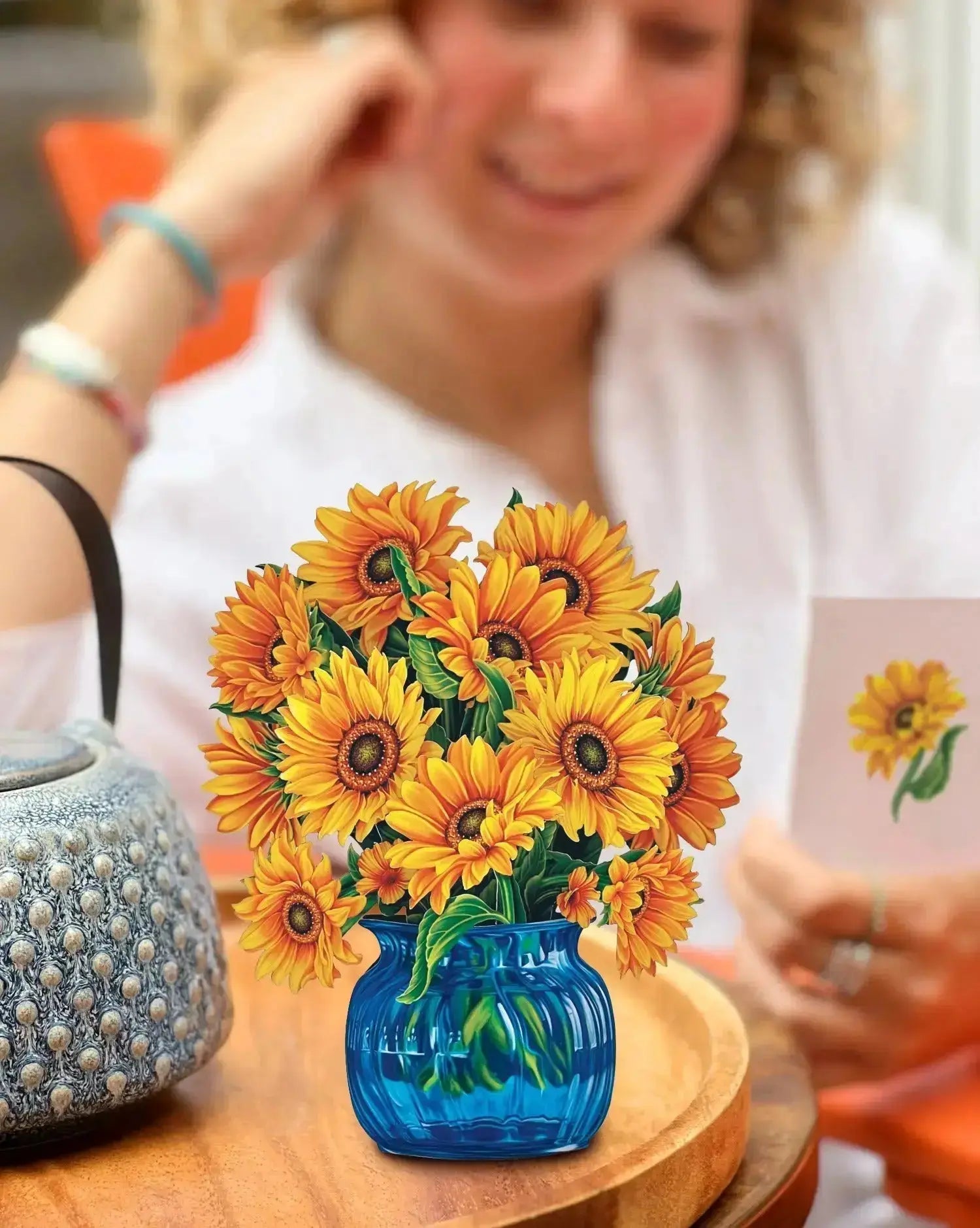 A person with curly hair, seated at an orange table, smiles while holding a card featuring a sunflower. In the foreground is a blue glass vase brimming with Freshcut Paper's Mini Sunflowers that seem almost like Van Gogh’s sunflowers. On the table, there's also a textured blue handbag.