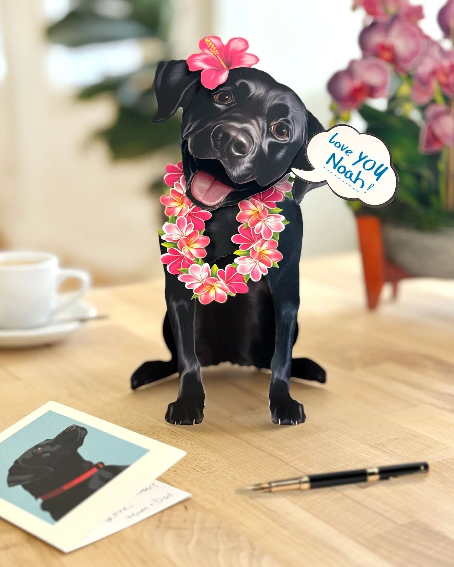 Freshcut Paper Black Lab pop-up card with pink flower leis on a wooden surface with a card and pen.