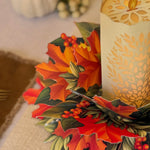 An autumn-themed 3D pop-up paper centerpiece featuring colorful maple leaves, holly, berries, and pinecones, styled next to a lit cream-colored pillar candle with a laser-cut leaf pattern.