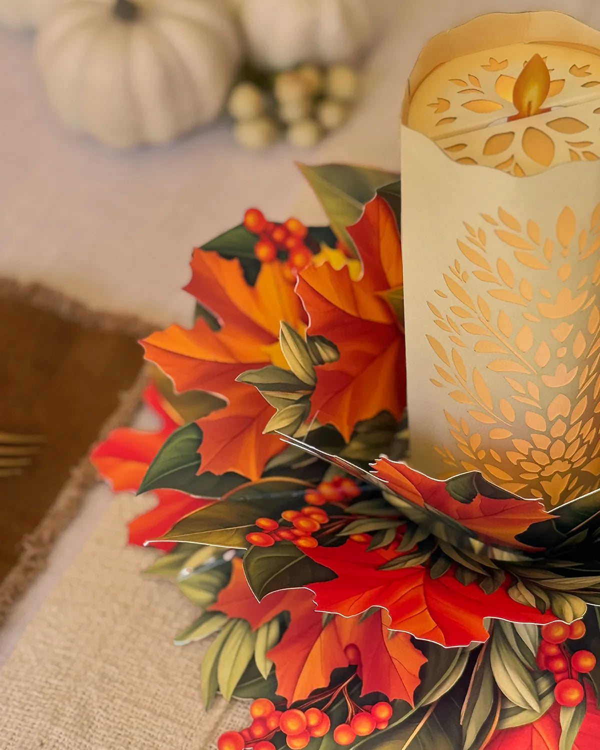 An autumn-themed 3D pop-up paper centerpiece featuring colorful maple leaves, holly, berries, and pinecones, styled next to a lit cream-colored pillar candle with a laser-cut leaf pattern.