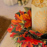 An autumn-themed 3D pop-up paper centerpiece featuring colorful maple leaves, holly, berries, and pinecones, styled next to a lit cream-colored pillar candle with a laser-cut leaf pattern.