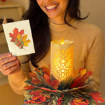A woman in a beige sweater holds a small printed card showing an autumn leaf design next to a glowing LED candle holder made of layered paper leaves in red, orange, and green.