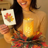 A woman in a beige sweater holds a small printed card showing an autumn leaf design next to a glowing LED candle holder made of layered paper leaves in red, orange, and green.