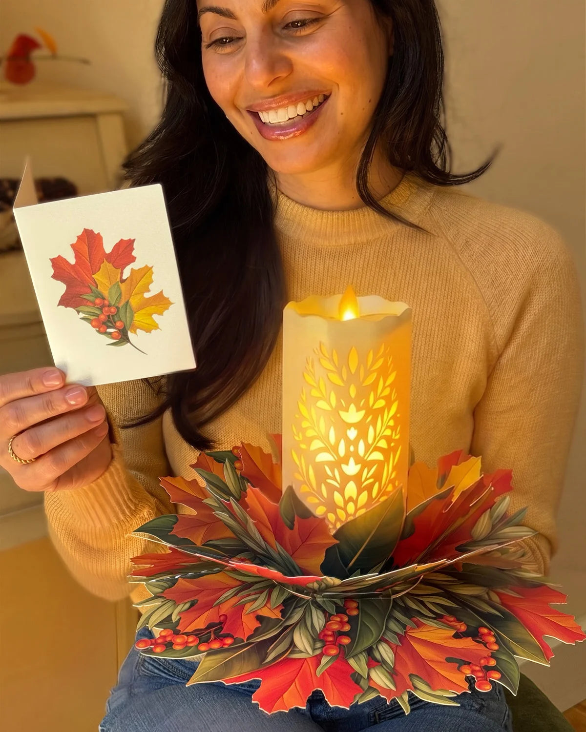A woman in a beige sweater holds a small printed card showing an autumn leaf design next to a glowing LED candle holder made of layered paper leaves in red, orange, and green.