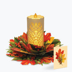 An autumn-themed decorative centerpiece featuring a glowing pillar candle with a leaf cutout pattern, surrounded by a ring of red and orange maple leaves, green foliage, and small red berries, with a matching small card standing beside it on a light gray background.