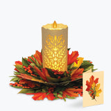 An autumn-themed decorative centerpiece featuring a glowing pillar candle with a leaf cutout pattern, surrounded by a ring of red and orange maple leaves, green foliage, and small red berries, with a matching small card standing beside it on a light gray background.