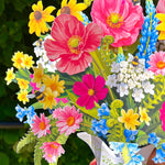 A close-up of a colorful bouquet of artificial flowers arranged in a white pitcher vase, held by a hand against a blurred green outdoor background.