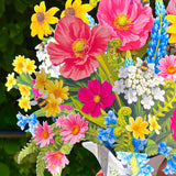 A close-up of a colorful bouquet of artificial flowers arranged in a white pitcher vase, held by a hand against a blurred green outdoor background.