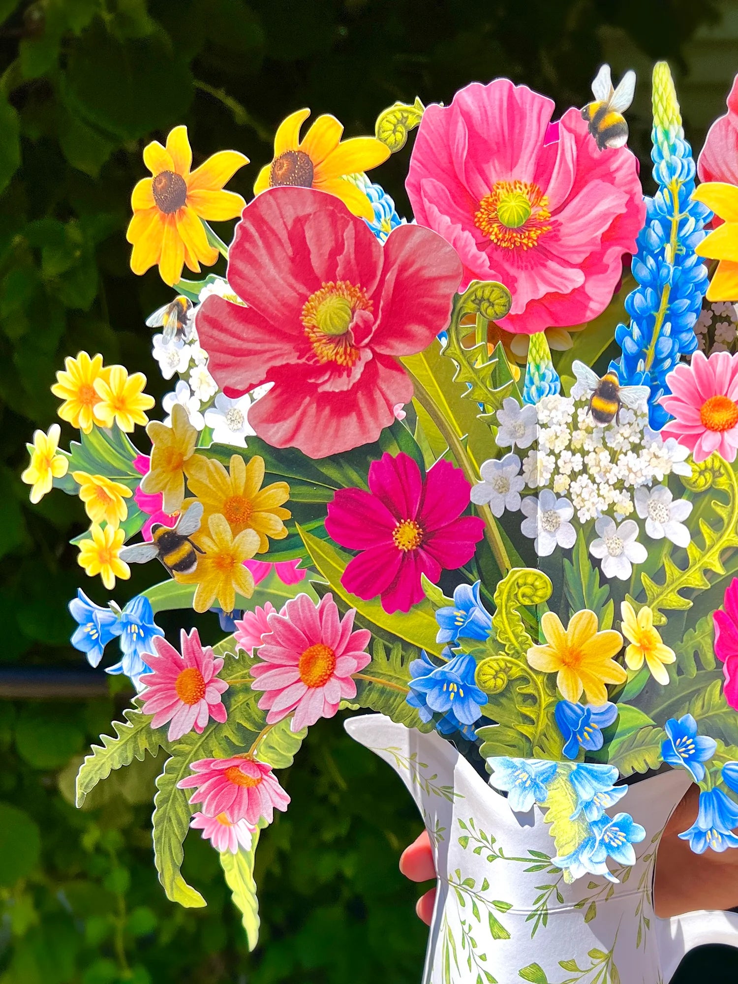 A close-up of a colorful bouquet of artificial flowers arranged in a white pitcher vase, held by a hand against a blurred green outdoor background.
