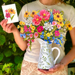 A woman outdoors holds a white ceramic pitcher filled with a colorful bouquet of flowers, while also holding a small card with a floral illustration. The background is lush green foliage.