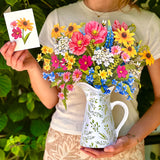 A woman outdoors holds a white ceramic pitcher filled with a colorful bouquet of flowers, while also holding a small card with a floral illustration. The background is lush green foliage.