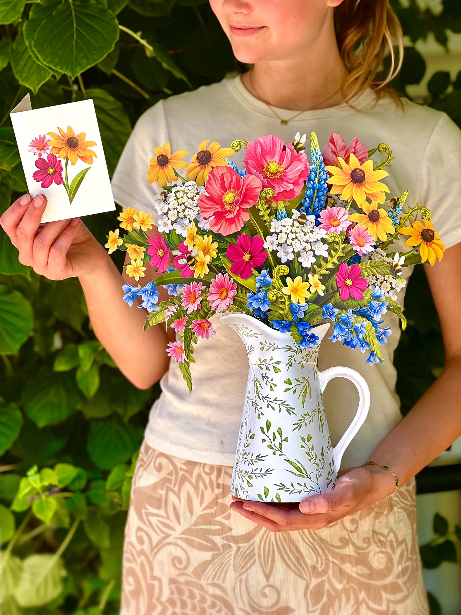 A woman outdoors holds a white ceramic pitcher filled with a colorful bouquet of flowers, while also holding a small card with a floral illustration. The background is lush green foliage.