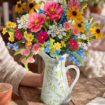 A colorful wildflower bouquet arranged in a white ceramic pitcher with green leaf patterns, set on a rustic wooden table with a person's arm visible holding the pitcher.