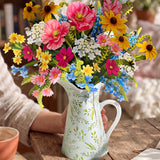 A colorful wildflower bouquet arranged in a white ceramic pitcher with green leaf patterns, set on a rustic wooden table with a person's arm visible holding the pitcher.
