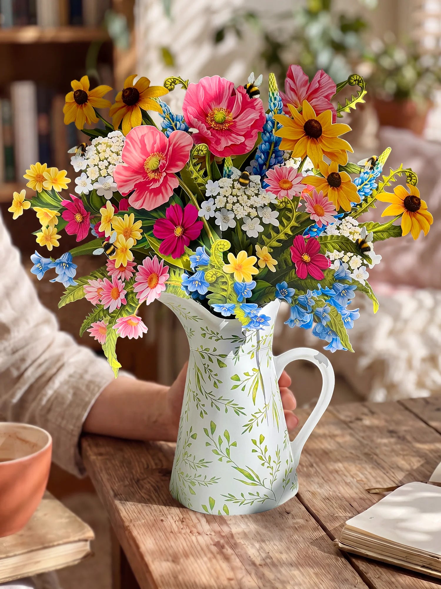 A colorful wildflower bouquet arranged in a white ceramic pitcher with green leaf patterns, set on a rustic wooden table with a person's arm visible holding the pitcher.