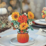 The Mini Pumpkin Harvest by Freshcut Paper is a festive autumn centerpiece showcasing an arrangement of vibrant fall flowers in shades of orange, yellow, and white, including sunflowers and dahlias. These beautiful blooms are nestled in an orange vase shaped like a pumpkin and rest on a white plate as part of a table setting with blurred lights in the background.