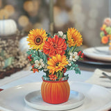 The Mini Pumpkin Harvest by Freshcut Paper is a festive autumn centerpiece showcasing an arrangement of vibrant fall flowers in shades of orange, yellow, and white, including sunflowers and dahlias. These beautiful blooms are nestled in an orange vase shaped like a pumpkin and rest on a white plate as part of a table setting with blurred lights in the background.