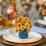 A table is set with white and brown plates, a folded cloth napkin, and a blue glass vase filled with vibrant Freshcut Paper Mini Sunflowers. In the background, there are more floral arrangements and blurred lights creating a warm, festive atmosphere.