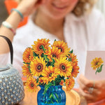 A person with curly hair, seated at an orange table, smiles while holding a card featuring a sunflower. In the foreground is a blue glass vase brimming with Freshcut Paper's Mini Sunflowers that seem almost like Van Gogh’s sunflowers. On the table, there's also a textured blue handbag.