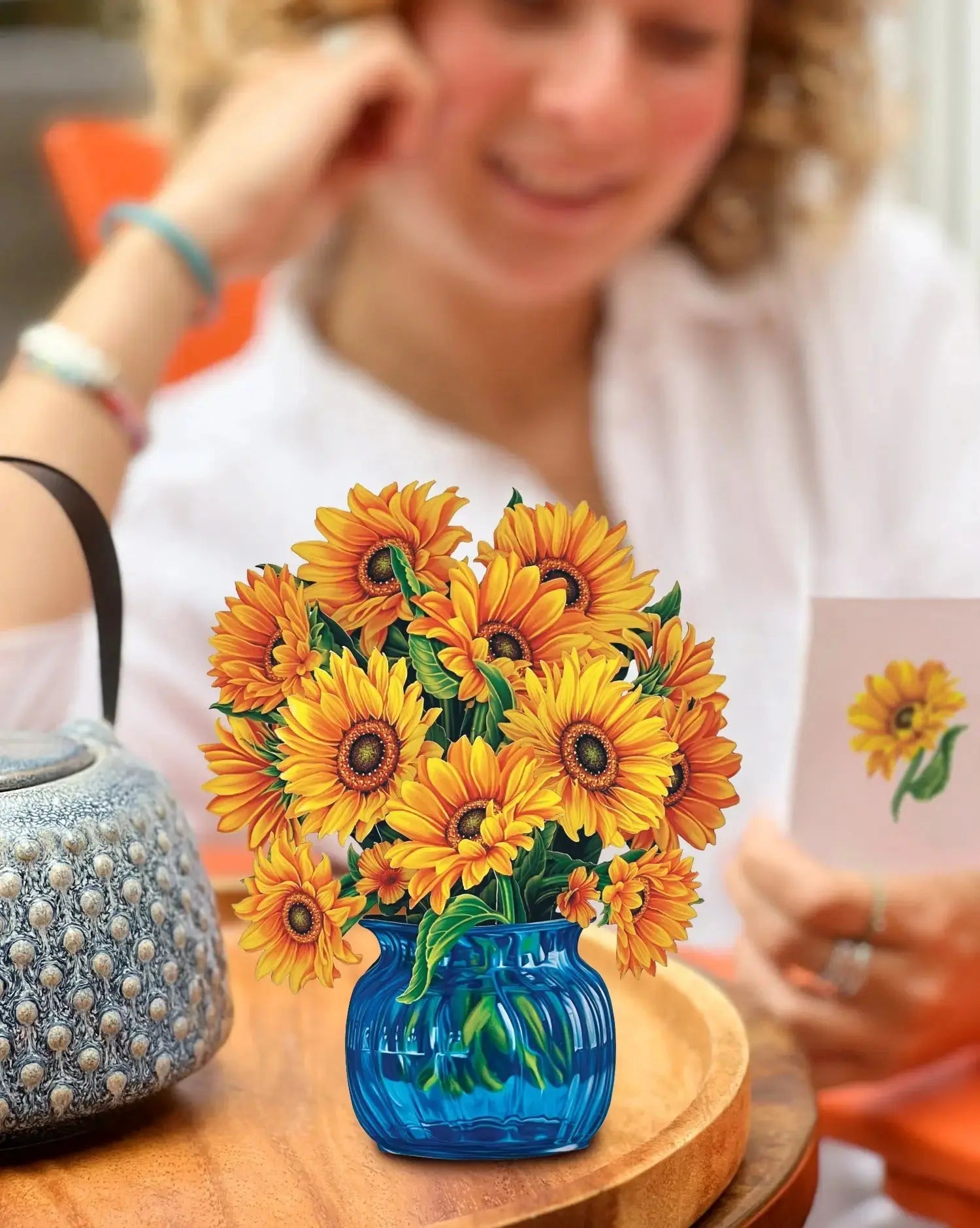 A person with curly hair, seated at an orange table, smiles while holding a card featuring a sunflower. In the foreground is a blue glass vase brimming with Freshcut Paper's Mini Sunflowers that seem almost like Van Gogh’s sunflowers. On the table, there's also a textured blue handbag.