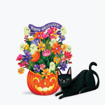A festive Halloween illustration featuring a carved orange pumpkin filled with colorful flowers, small ghost figures, and a purple 'Happy Halloween!' banner, next to a paper black cat on a light gray background.