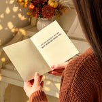 A person in a rust-brown knit sweater holds an open cream-colored book or journal in a warm, sunlit indoor setting. The pages show the text 'HOW YOUR HEART WANTS TO BE TOLD! READ IT.