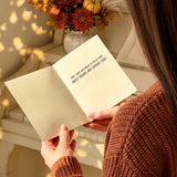 A person in a rust-brown knit sweater holds an open cream-colored book or journal in a warm, sunlit indoor setting. The pages show the text 'HOW YOUR HEART WANTS TO BE TOLD! READ IT.