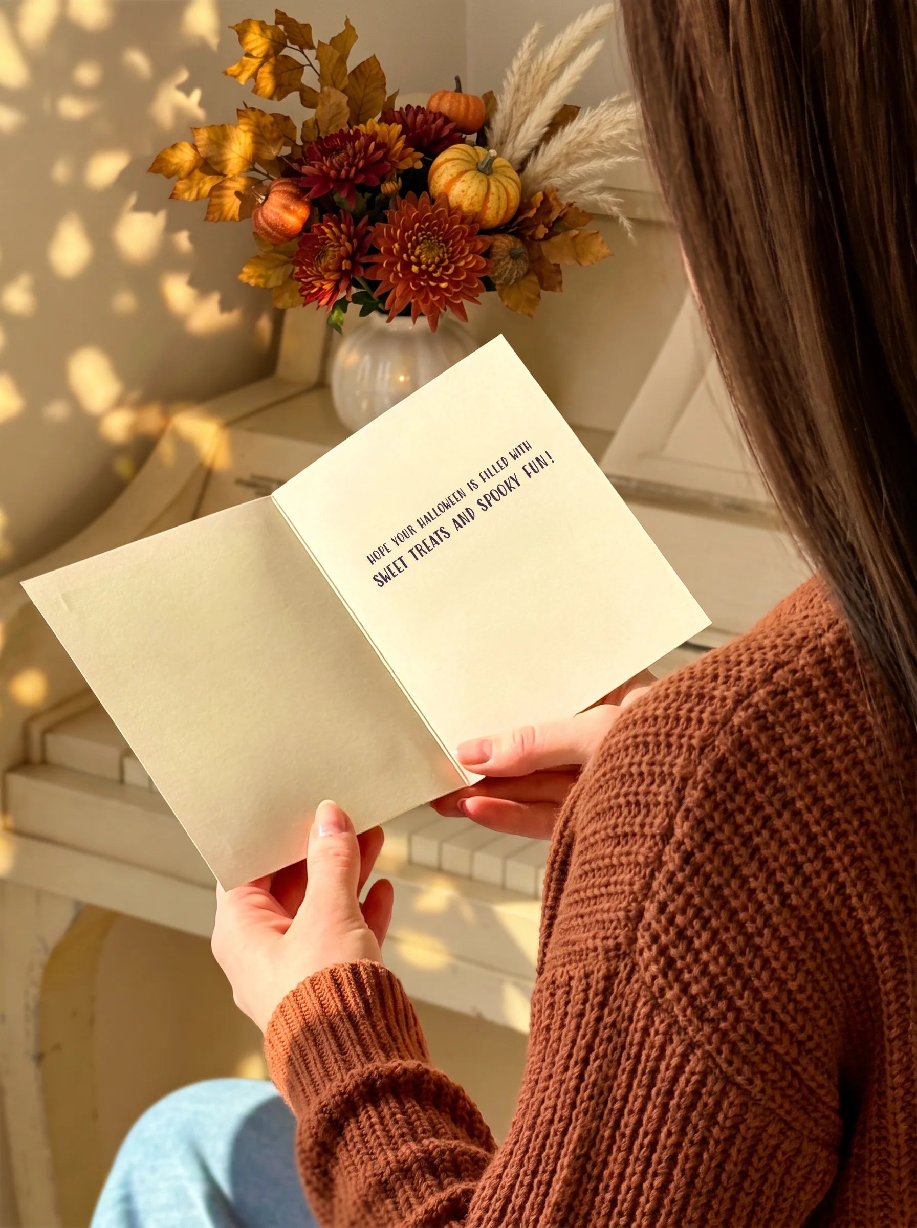 A person in a rust-brown knit sweater holds an open cream-colored book or journal in a warm, sunlit indoor setting. The pages show the text 'HOW YOUR HEART WANTS TO BE TOLD! READ IT.