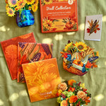 A styled flat-lay of fall pop-up cards and decorative autumn-themed items arranged on a green fabric background with dappled sunlight. Visible products include a boxed set of 3 fall-themed pop-up cards, several orange floral and corn patterned branded envelopes, a blue vase with yellow flowers, a small basket with colorful autumn decorations and flowers, and a white pumpkin vase with mixed flowers.