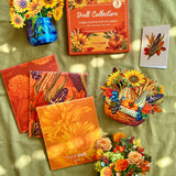 A styled flat-lay of fall pop-up cards and decorative autumn-themed items arranged on a green fabric background with dappled sunlight. Visible products include a boxed set of 3 fall-themed pop-up cards, several orange floral and corn patterned branded envelopes, a blue vase with yellow flowers, a small basket with colorful autumn decorations and flowers, and a white pumpkin vase with mixed flowers.