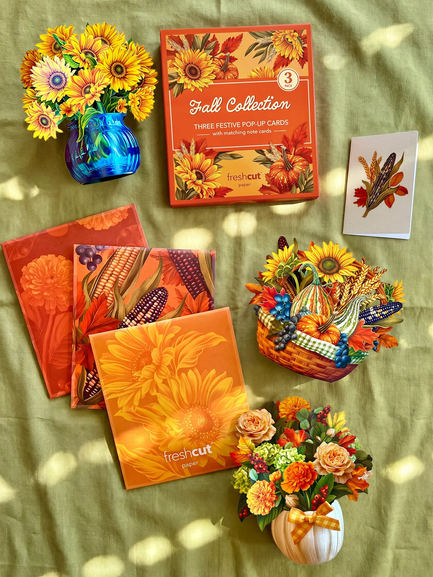 A styled flat-lay of fall pop-up cards and decorative autumn-themed items arranged on a green fabric background with dappled sunlight. Visible products include a boxed set of 3 fall-themed pop-up cards, several orange floral and corn patterned branded envelopes, a blue vase with yellow flowers, a small basket with colorful autumn decorations and flowers, and a white pumpkin vase with mixed flowers.