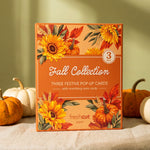 A boxed 3-card set labeled 'Fall Collection' stands upright on a draped fabric surface with pumpkins around it. The box has autumn floral artwork with sunflowers, pumpkins, and leaves, and the visible brand 'freshcut paper' appears near the bottom.