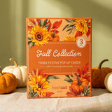 A boxed 3-card set labeled 'Fall Collection' stands upright on a draped fabric surface with pumpkins around it. The box has autumn floral artwork with sunflowers, pumpkins, and leaves, and the visible brand 'freshcut paper' appears near the bottom.