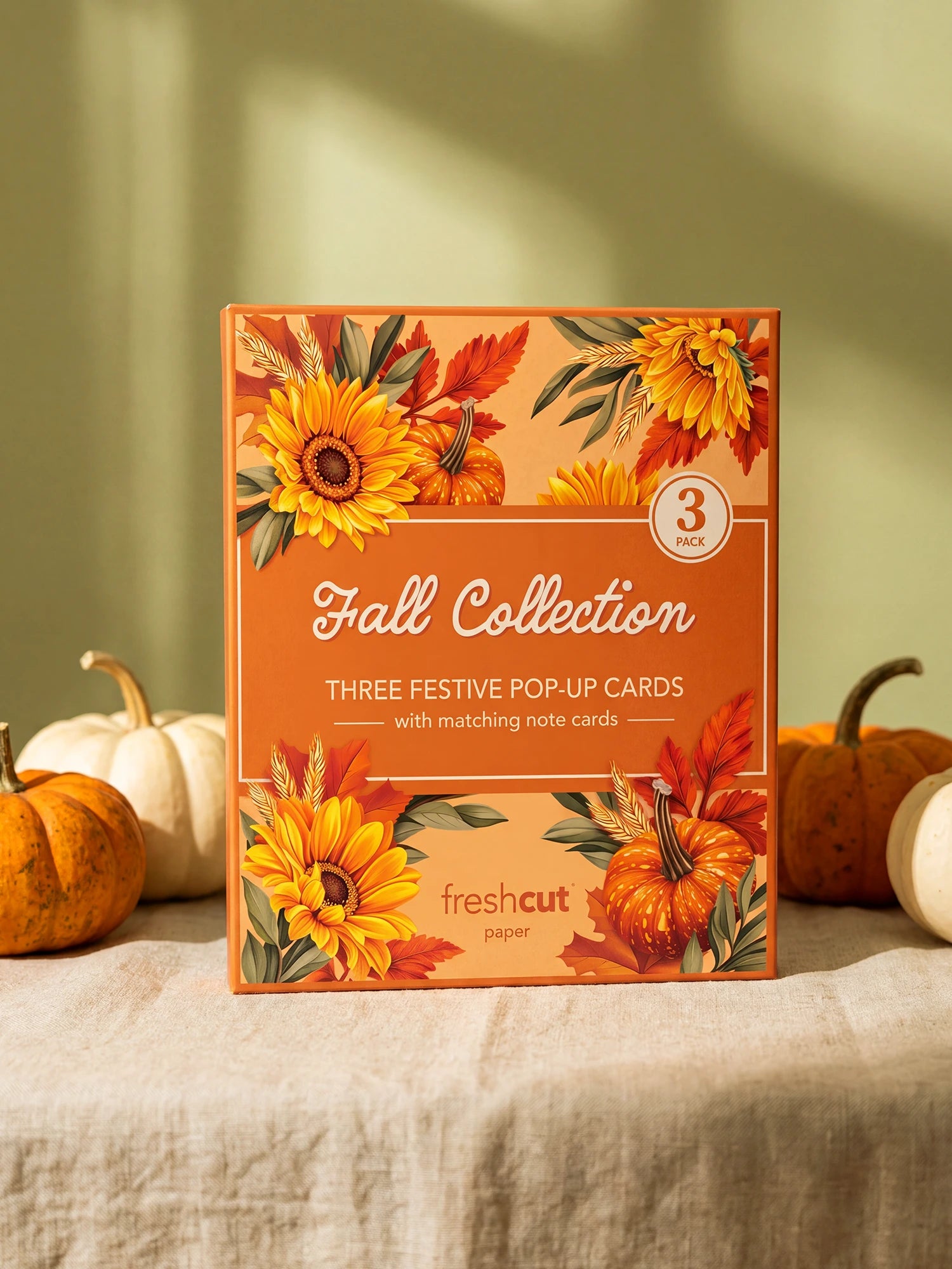 A boxed 3-card set labeled 'Fall Collection' stands upright on a draped fabric surface with pumpkins around it. The box has autumn floral artwork with sunflowers, pumpkins, and leaves, and the visible brand 'freshcut paper' appears near the bottom.