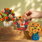 Three decorative paper flower arrangements displayed on rustic wooden steps against a beige wall with leaf shadows. A human hand is reaching toward the center arrangement, which is a small autumn-themed basket with pumpkins, sunflowers, and berries.