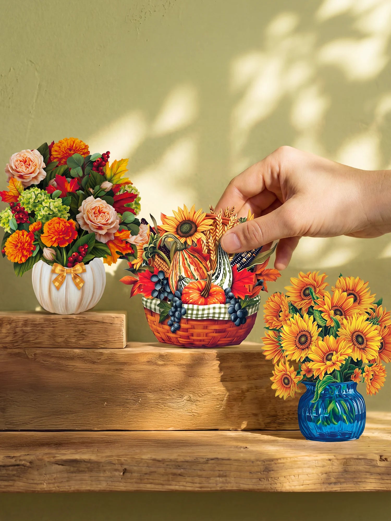 Three decorative paper flower arrangements displayed on rustic wooden steps against a beige wall with leaf shadows. A human hand is reaching toward the center arrangement, which is a small autumn-themed basket with pumpkins, sunflowers, and berries.