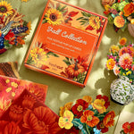 A styled flat-lay of a the 'Fall Collection' boxed set of five matching pop-up cards in an orange box, surrounded by colorful artificial flowers and autumn-themed packaging on a light green surface.
