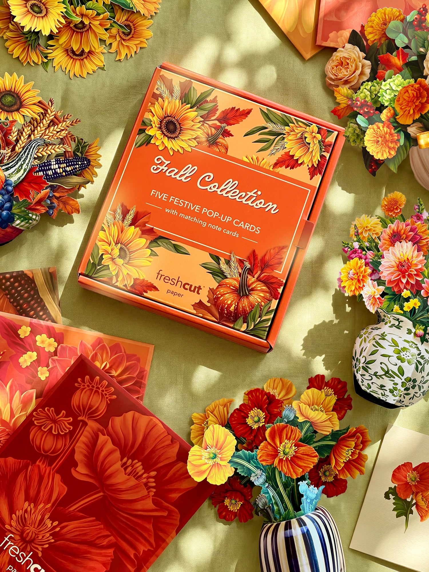 A styled flat-lay of a the 'Fall Collection' boxed set of five matching pop-up cards in an orange box, surrounded by colorful artificial flowers and autumn-themed packaging on a light green surface.