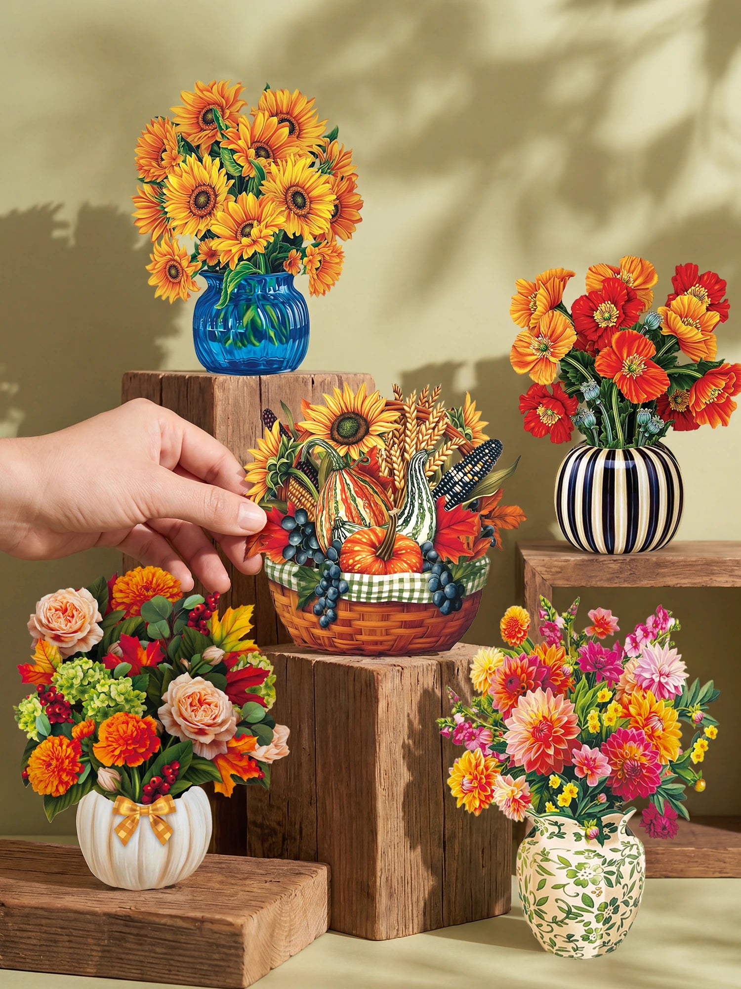 A hand holds a small woven basket filled with autumn flowers and decorative stems, surrounded by several other floral arrangements in colorful vases and pots on wooden blocks against a warm sage green wall.