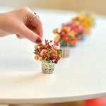 A close-up of a hand placing a tiny miniature dried flower bouquet in a small textured glass pot on a white table, with a row of similar miniature flower arrangements blurred in the background.