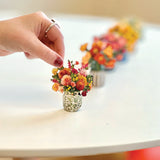 A close-up of a hand placing a tiny miniature dried flower bouquet in a small textured glass pot on a white table, with a row of similar miniature flower arrangements blurred in the background.