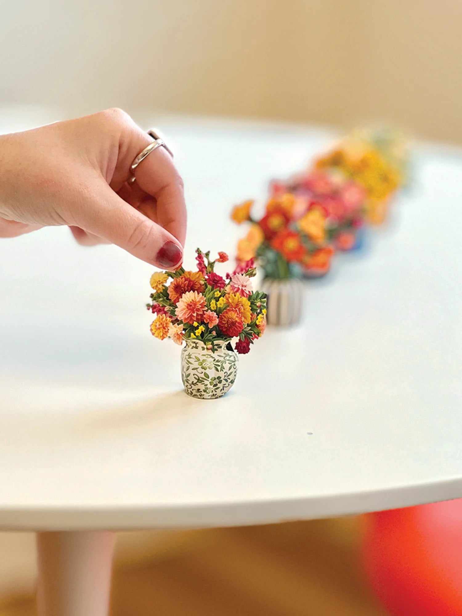 A close-up of a hand placing a tiny miniature dried flower bouquet in a small textured glass pot on a white table, with a row of similar miniature flower arrangements blurred in the background.
