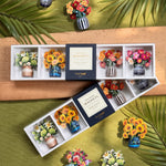 A floral magnet set displayed in two open white trays. The set includes miniature artificial flower bouquets in colorful vases and decorative striped and patterned ceramic vases, arranged on a wooden board over a green textured background with palm leaves.