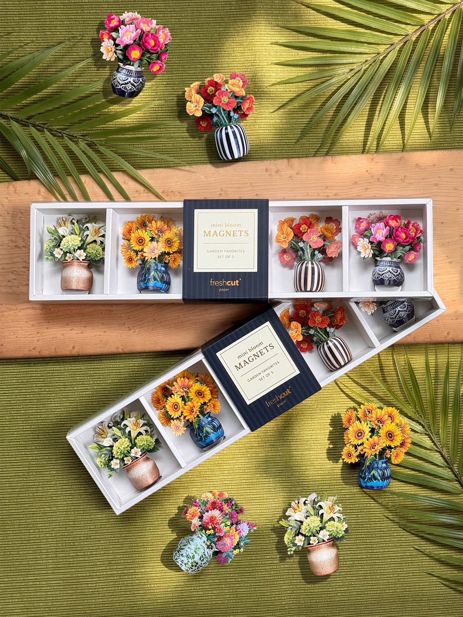 A floral magnet set displayed in two open white trays. The set includes miniature artificial flower bouquets in colorful vases and decorative striped and patterned ceramic vases, arranged on a wooden board over a green textured background with palm leaves.