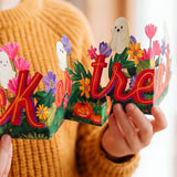 A person in a mustard yellow knit sweater holds a colorful Halloween-themed decorative letter-form sign with the words "trick or treat" in red letters, surrounded by flowers, ghosts, pumpkins, and green leaves.