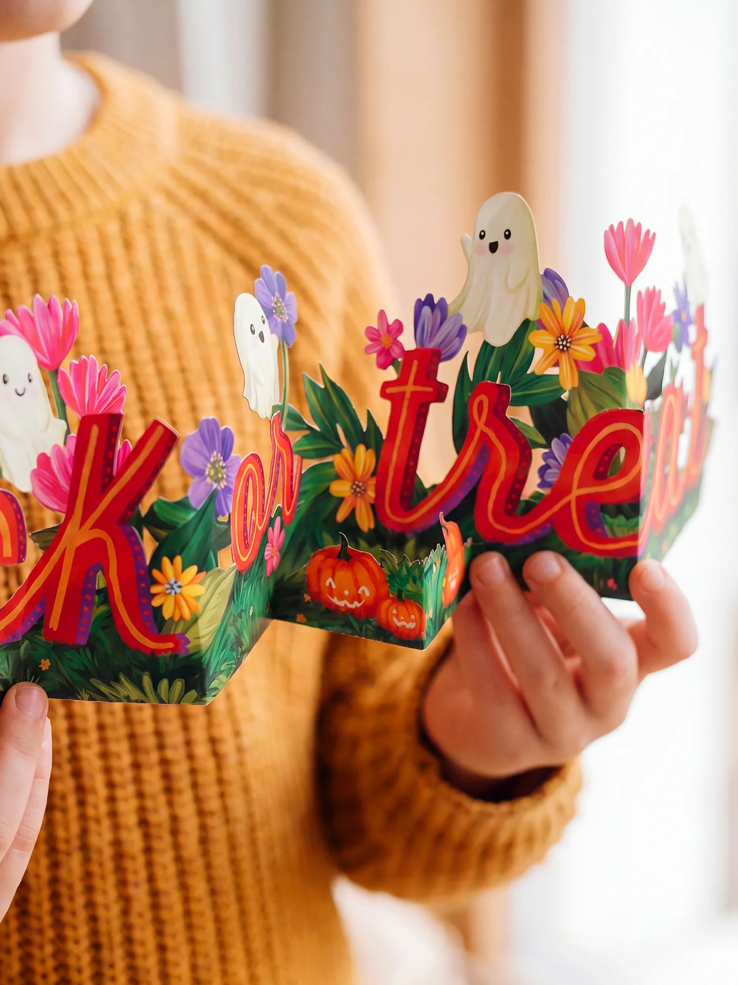 A person in a mustard yellow knit sweater holds a colorful Halloween-themed decorative letter-form sign with the words "trick or treat" in red letters, surrounded by flowers, ghosts, pumpkins, and green leaves.