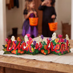 A Halloween-themed decorative tabletop sign reading 'trick or treat' with colorful flowers, pumpkins, ghosts, and green leaves, placed on a wooden table in a home setting. Two children in witch and fairy costumes are blurred in the background holding orange trick-or-treat buckets.