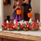 A Halloween-themed decorative tabletop sign reading 'trick or treat' with colorful flowers, pumpkins, ghosts, and green leaves, placed on a wooden table in a home setting. Two children in witch and fairy costumes are blurred in the background holding orange trick-or-treat buckets.