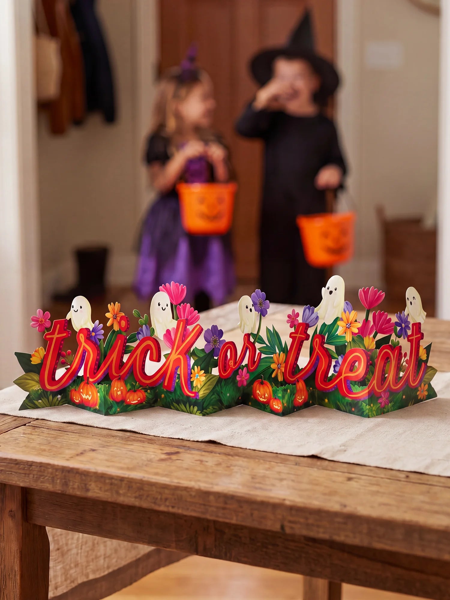 A Halloween-themed decorative tabletop sign reading 'trick or treat' with colorful flowers, pumpkins, ghosts, and green leaves, placed on a wooden table in a home setting. Two children in witch and fairy costumes are blurred in the background holding orange trick-or-treat buckets.